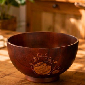 May include: A dark brown wooden bowl featuring a laser-engraved illustration of a large, round, cat-like creature with pointed ears and whiskers, accompanied by a smaller version of the same creature, centered on the exterior surface.