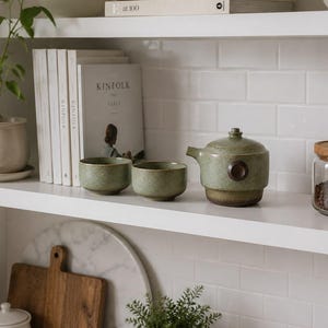 Może przedstawiać: Ceramiczny zestaw do herbaty składający się z małego czajniczka z ciemnobrązowym okrągłym uchwytem bocznym oraz dwóch pasujących czarek bez uszek. Zestaw posiada nakrapianą szałwiowo-zieloną glazurę z ciemnobrązową podstawą.