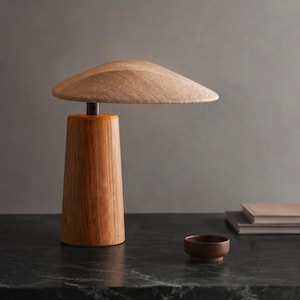May include: A table lamp featuring a cylindrical wooden base with a visible grain pattern and a wide, shallow, mushroom-shaped shade made of a textured, light-colored material, connected by a dark metal stem.