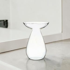 May include: A clear glass vase with a teardrop-shaped base and a wide, flared rim. The vessel features a smooth, transparent surface and a narrow neck that transitions into a rounded, bulbous bottom, creating a simple and functional silhouette.