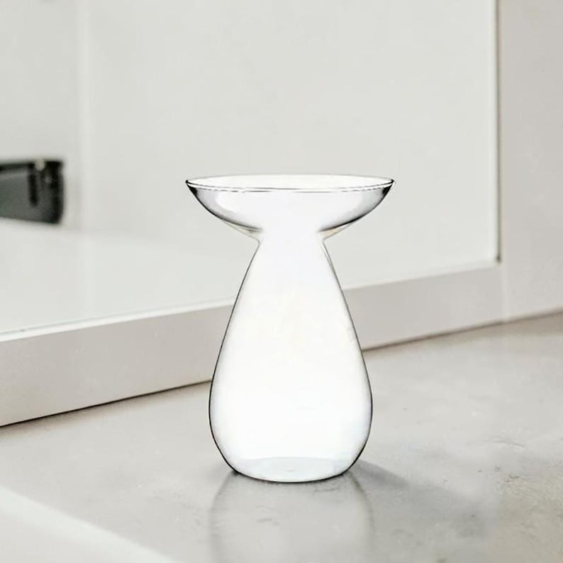 May include: A clear glass vase with a teardrop-shaped base and a wide, flared rim. The vessel features a smooth, transparent surface and a narrow neck that transitions into a rounded, bulbous bottom, creating a simple and functional silhouette.