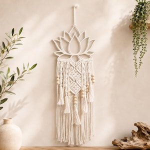 May include: A wall hanging featuring a white lotus flower cutout at the top, connected to a cream-colored macrame section with intricate knotted patterns, wooden beads, and long fringe tassels hanging from the bottom of the fiber design.
