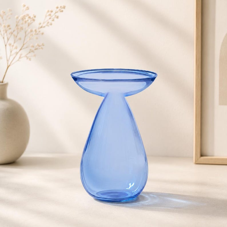 May include: A translucent blue glass vase with a teardrop-shaped body and a wide, flat, circular rim. The vase features a smooth, polished surface and a narrow neck that transitions into a rounded base, creating a distinct silhouette.