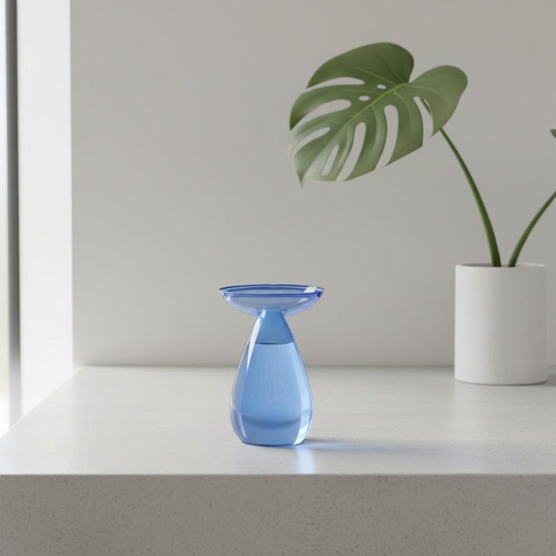 May include: A small, translucent blue glass vase with a teardrop-shaped base and a wide, flat, circular rim. The vase has a smooth, polished surface and a clear, hollow interior, reflecting light through its vibrant, cool-toned blue glass structure.