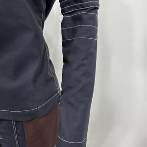 May include: A long-sleeved black jacket featuring prominent white contrast stitching along the seams and three horizontal white stitched bands around the upper arm. The garment includes a dark maroon panel on the lower side section.