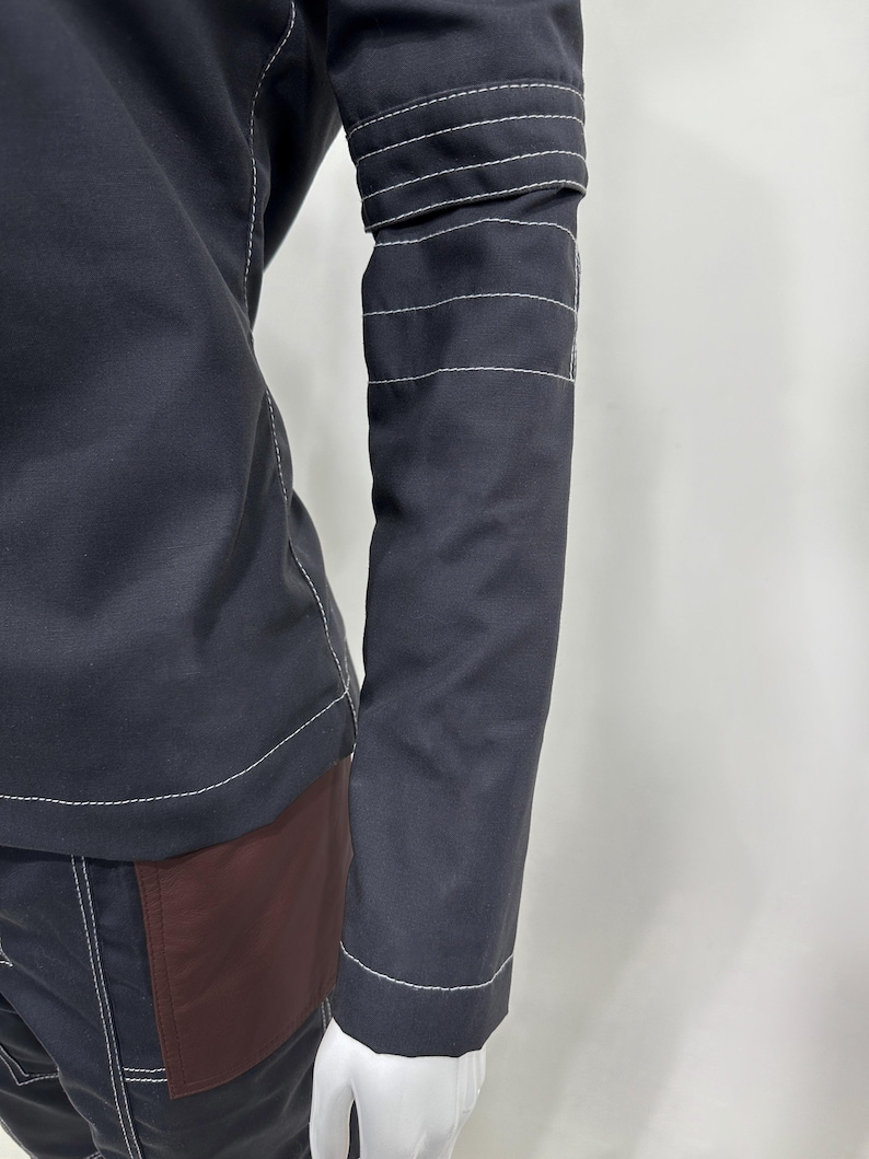 May include: A long-sleeved black jacket featuring prominent white contrast stitching along the seams and three horizontal white stitched bands around the upper arm. The garment includes a dark maroon panel on the lower side section.