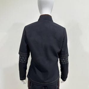 May include: A dark charcoal jacket with a high collar and brown trim, featuring white contrast stitching and a geometric grid pattern on the sleeves. The jacket is paired with matching dark charcoal trousers that also display white contrast stitching.