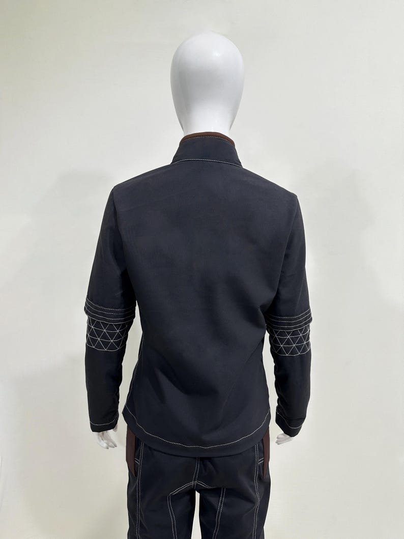 May include: A dark charcoal jacket with a high collar and brown trim, featuring white contrast stitching and a geometric grid pattern on the sleeves. The jacket is paired with matching dark charcoal trousers that also display white contrast stitching.