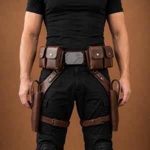 May include: A brown leather utility belt featuring four rectangular pouches with silver-toned snap closures, a central metallic buckle with geometric detailing, and two integrated holsters secured by adjustable leg straps over black trousers.