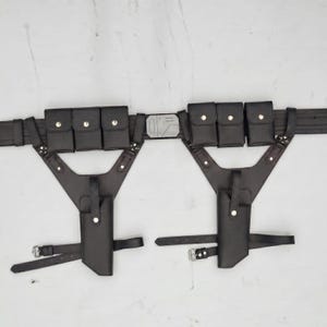 May include: A black utility belt featuring two symmetrical holster attachments, each equipped with three small rectangular pouches with snap closures and a vertical holster sleeve. The central buckle is a rectangular silver-toned metal piece.