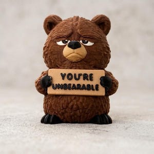 Könnte beinhalten: Eine braune Bärenfigur mit strukturierter Felloptik, die ein beigefarbenes rechteckiges Schild mit dem schwarzen Text "YOU'RE UNBEARABLE" hält. Der Bär hat einen mürrischen Gesichtsausdruck mit zusammengekniffenen Augen, einer schwarzen Nase und schwarzen Pfoten.