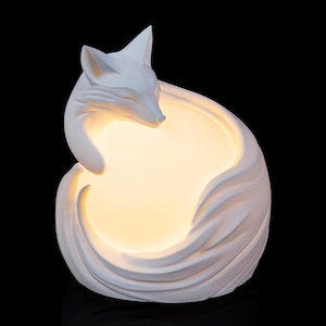 May include: A white, fox-shaped lamp with a smooth, matte finish. The fox is curled into a circular shape, with its head resting near its tail. The center of the fox's body emits a warm, soft, glowing light, creating a bright, circular illumination.