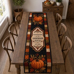 May include: A rectangular fabric table runner featuring a black center panel with orange pumpkins, autumn leaves, and a central cream-colored plaque printed with the text Thanksgiving at the Anderson House Est. 1994, bordered by a patchwork of fall-colored squares.