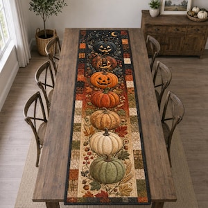 Op de afbeelding: Een lange rechthoekige tafelloper met een verticale rangschikking van zeven pompoenen in verschillende kleuren en texturen, waaronder een donkere Halloween-pompoen, oranje, beige, witte en groene pompoenen, omringd door herfstbladeren en eikels.