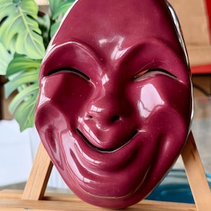 May include: A burgundy ceramic mask with a smiling face. The mask has closed eyes and a wide, upturned mouth. The surface is glossy and reflects light. The mask is mounted on a small wooden easel, suggesting it is a decorative item or artwork.
