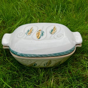 May include: A white ceramic casserole dish with a lid, featuring a leaf design in yellow, green, and blue. The dish has handles on each side and a green border. The casserole dish is on a bed of green grass.