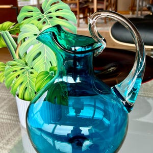 May include: A teal blue glass pitcher with a clear glass handle. The pitcher has a round body and a wide mouth.