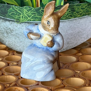 May include: A ceramic figurine of a brown rabbit wearing a light blue dress with a white collar. The rabbit is seated on a small chair, holding a small white teacup in one hand and a piece of yellow toast in the other, with a glossy finish.