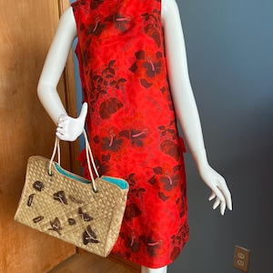 May include: A sleeveless red dress with a floral print and the word "Hawaii" repeated throughout. A woven straw handbag with brown embellishments and a turquoise interior is held by a mannequin.