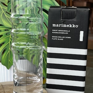 May include: A clear glass flute glass with a bamboo-like design stands next to a black and white striped box. The box features the brand name "marimekko" and text in multiple languages. The glass is empty and reflects the surrounding greenery.