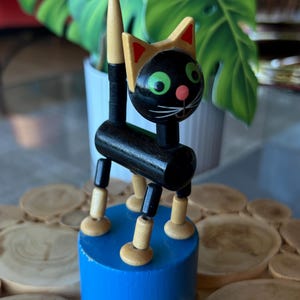 May include: A black wooden cat toy with a blue base. The cat has green eyes, a pink nose, and a yellow-trimmed head. Its legs and tail are made of wood and black joints. The toy is standing on a blue cylinder.