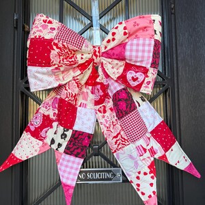 May include: A large, decorative fabric bow featuring a patchwork design in various shades of red, pink, and white. The fabric squares display diverse patterns including hearts, gingham, stripes, and floral motifs, creating a vibrant, quilted appearance.