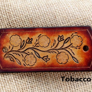 May include: A rectangular leather hair barrette with a floral design in brown and black. The patch is embossed with a vine of flowers and leaves. The text "Tobacco" is printed below the patch.