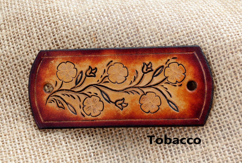 May include: A rectangular leather hair barrette with a floral design in brown and black. The patch is embossed with a vine of flowers and leaves. The text "Tobacco" is printed below the patch.