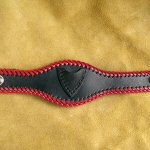 Peut inclure: Un bracelet en cuir noir avec des coutures rouges et deux boutons-pression argentés. Le bracelet a une petite poche triangulaire sur le devant.