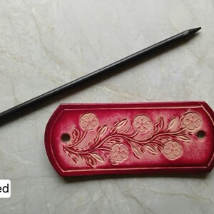 May include: A red leather tag with a floral design and two holes, next to a black pencil. The tag features embossed flowers and leaves. The word "Red" is in a white box in the lower left corner.