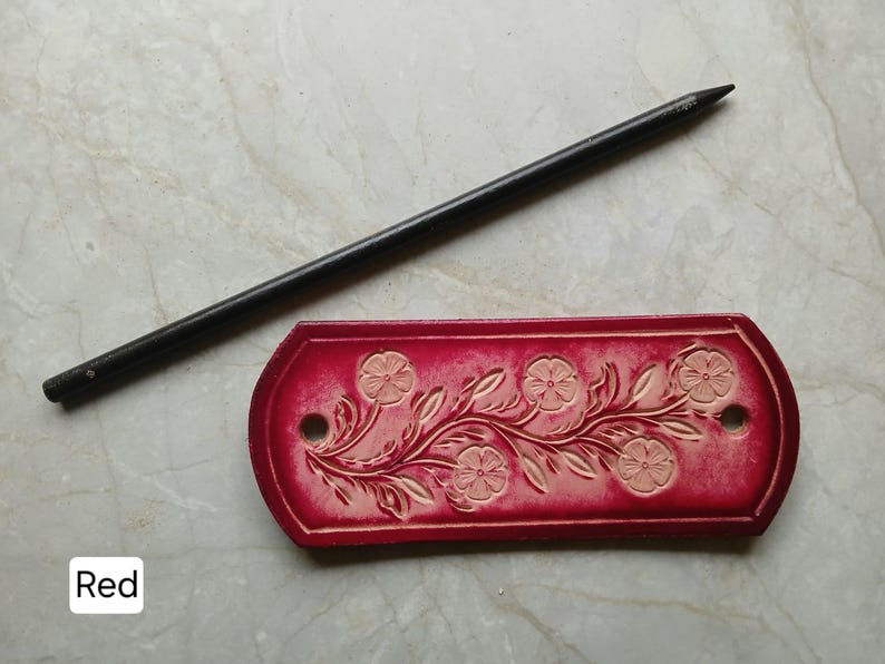 May include: A red leather tag with a floral design and two holes, next to a black pencil. The tag features embossed flowers and leaves. The word "Red" is in a white box in the lower left corner.