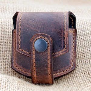 May include: A dark brown leather pouch with a flap closure secured by a single matte metal snap button. The pouch features visible stitching along the edges and a distressed, textured surface finish, providing a structured and compact appearance.