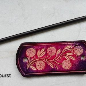May include: A handcrafted leather hair stick and barrette set. The barrette is a deep purple with a floral design and the text "Purpleburst". The hair stick is black.