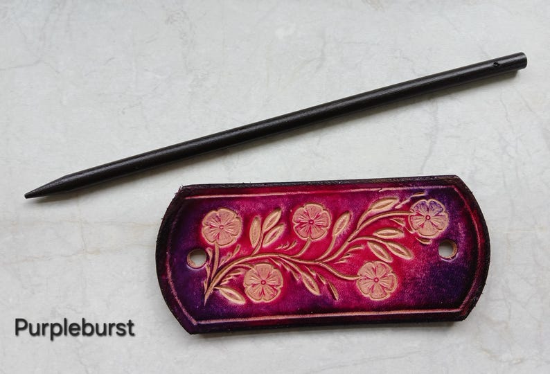 May include: A handcrafted leather hair stick and barrette set. The barrette is a deep purple with a floral design and the text "Purpleburst". The hair stick is black.
