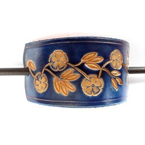 May include: A dark blue leather hair cuff with a floral design in a lighter shade of brown. A metal stick is inserted through the cuff. The cuff is a hair accessory.