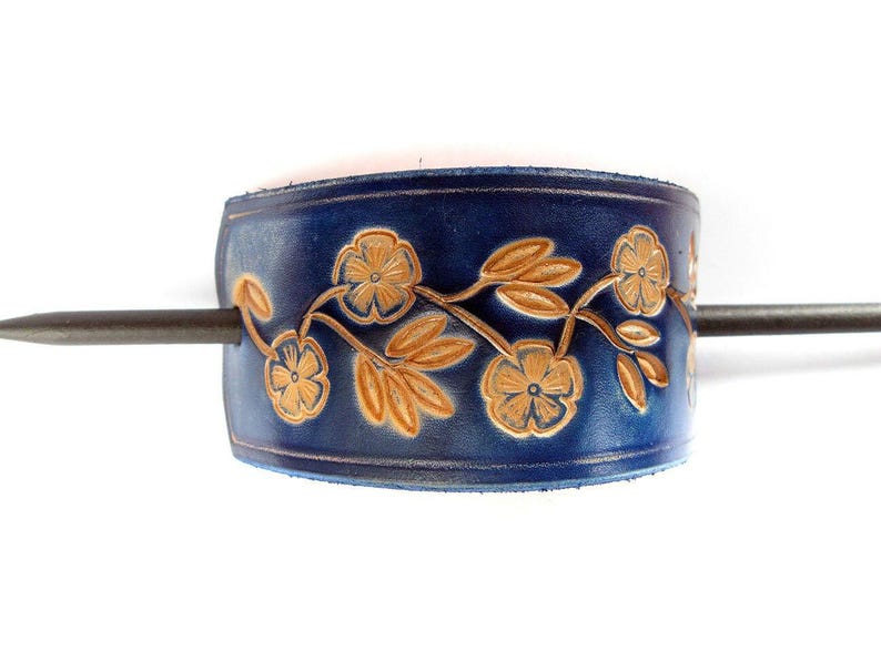 May include: A dark blue leather hair cuff with a floral design in a lighter shade of brown. A metal stick is inserted through the cuff. The cuff is a hair accessory.