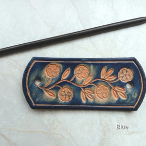 May include: A blue leather hair barrette with a floral design and a dark wooden hair stick. The barrette features embossed flowers and leaves in a lighter brown color. The word "Blue" is visible on the bottom right.