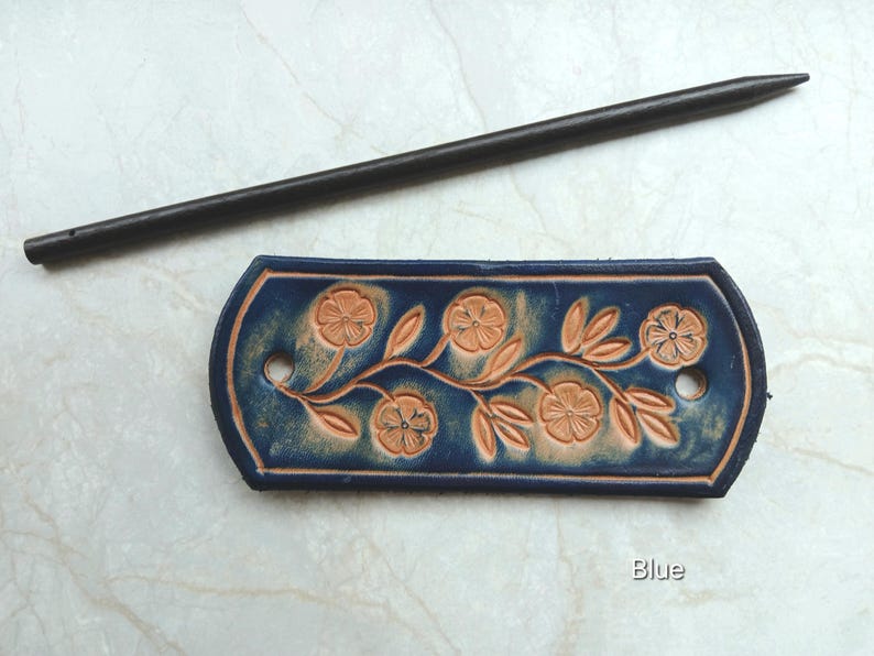 May include: A blue leather hair barrette with a floral design and a dark wooden hair stick. The barrette features embossed flowers and leaves in a lighter brown color. The word "Blue" is visible on the bottom right.