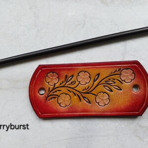 May include: A handcrafted leather hair slide with a floral design and a wooden stick. The hair accessory is red and orange with a floral pattern. The text "Cherryburst" is at the bottom of the image.