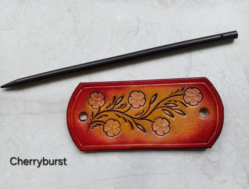 May include: A handcrafted leather hair slide with a floral design and a wooden stick. The hair accessory is red and orange with a floral pattern. The text "Cherryburst" is at the bottom of the image.