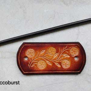 May include: A brown leather hair barrette with a floral design and a black wooden hair stick. The barrette is embossed with flowers and vines. The text "Tobacoburst" is at the bottom of the image.