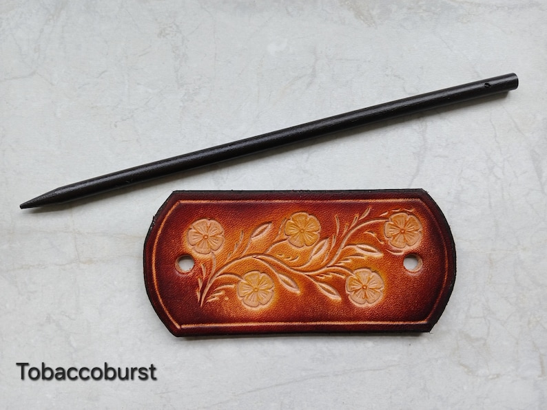 May include: A brown leather hair barrette with a floral design and a black wooden hair stick. The barrette is embossed with flowers and vines. The text "Tobacoburst" is at the bottom of the image.