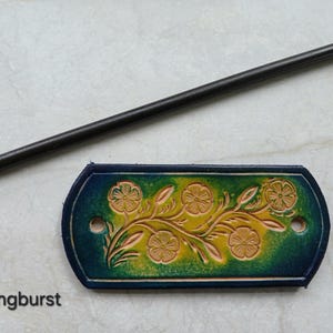 May include: A hand-tooled leather hair barrette with a floral design in shades of yellow and green, with a dark blue border. A long, black hair stick is positioned above the barrette. The word "Springburst" is printed below the barrette.