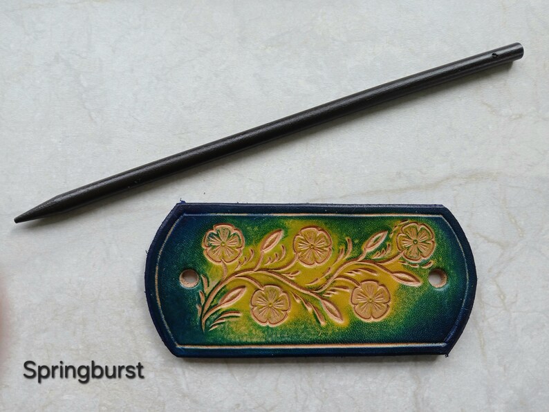 May include: A hand-tooled leather hair barrette with a floral design in shades of yellow and green, with a dark blue border. A long, black hair stick is positioned above the barrette. The word "Springburst" is printed below the barrette.
