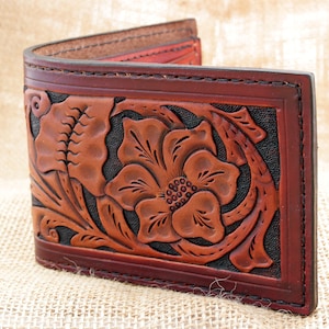 May include: A brown leather wallet with a floral design embossed on the front. The design features a large flower with intricate details and leaves.