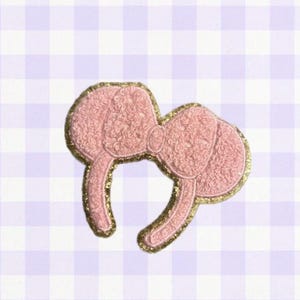 May include: An embroidered patch shaped like a headband with two round ears and a central bow. The design features a textured light pink surface with a shimmering gold border that outlines the entire shape of the ears, bow, and headband band.