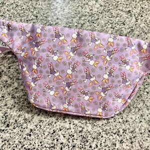 May include: A lavender fanny pack featuring an all-over print of Daisy Duck characters and small white daisies. The bag includes a matching light purple adjustable strap and a zipper closure along the top edge for secure storage of personal items.