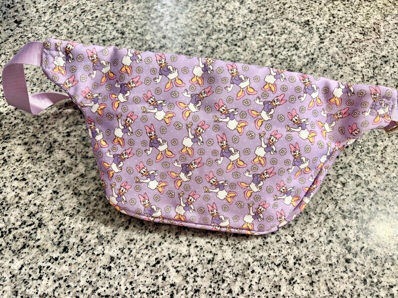 May include: A lavender fanny pack featuring an all-over print of Daisy Duck characters and small white daisies. The bag includes a matching light purple adjustable strap and a zipper closure along the top edge for secure storage of personal items.