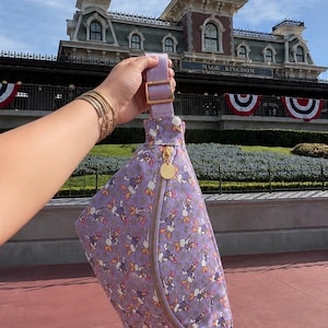 May include: A lavender fanny pack featuring an all-over print of Daisy Duck characters and small white flowers. The bag has a gold-tone zipper closure with a circular gold charm, an adjustable lavender strap, and a gold-tone metal buckle slider.