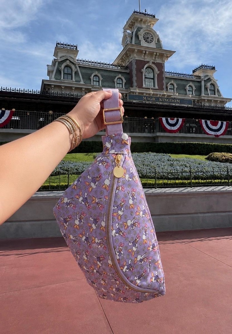 May include: A lavender fanny pack featuring an all-over print of Daisy Duck characters and small white flowers. The bag has a gold-tone zipper closure with a circular gold charm, an adjustable lavender strap, and a gold-tone metal buckle slider.